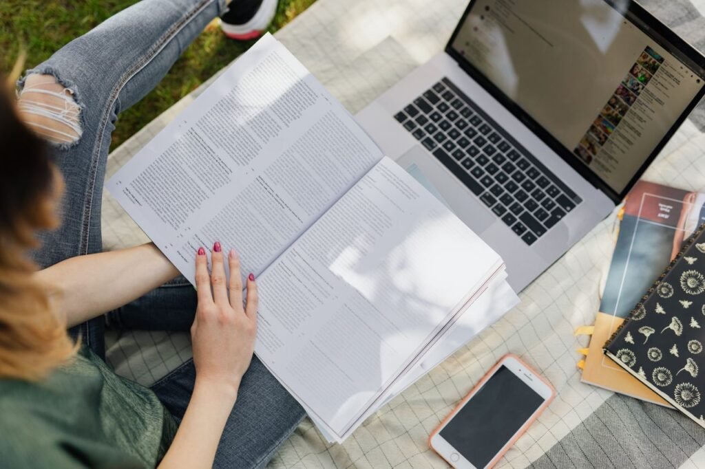 High angle of crop anonymous female student studying information in textbook while sitting with modern laptop and smartphone in sunny park