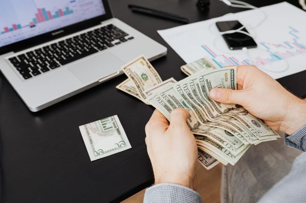 Person counting dollar bills at desk with laptop and financial charts, symbolizing finance management.