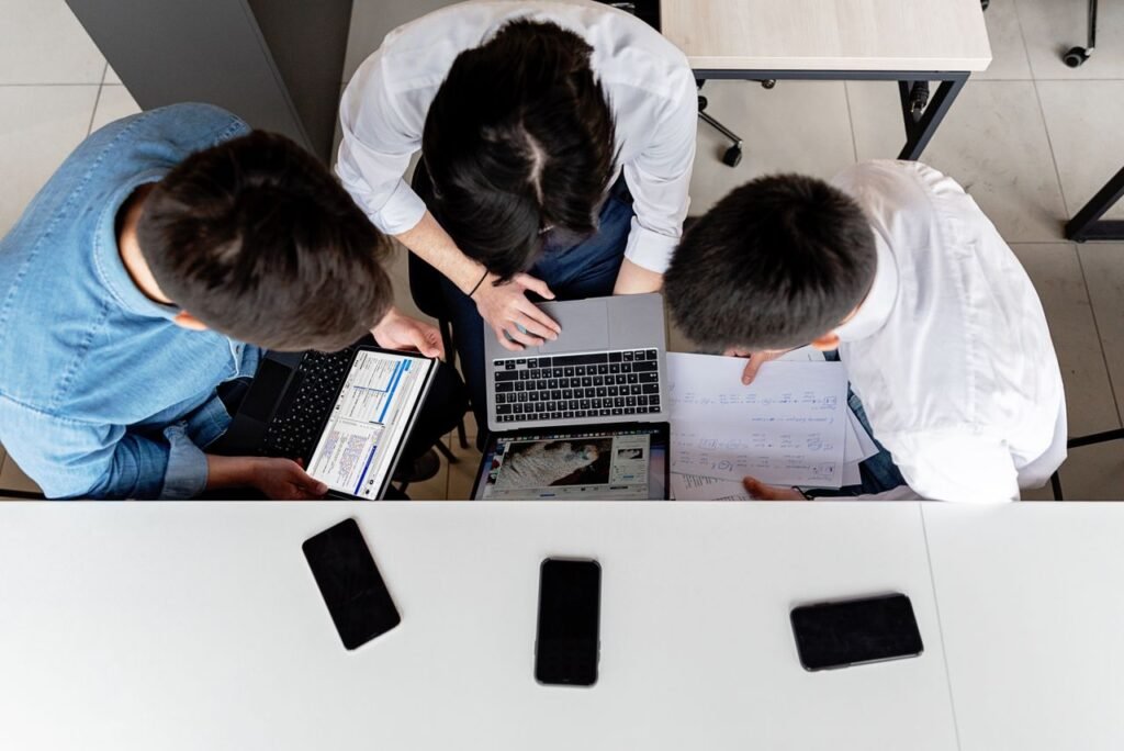 Three colleagues working together on laptops at an office desk with documents.