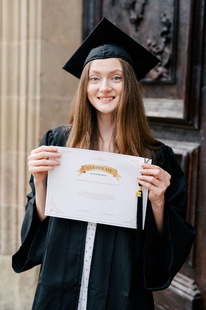 Happy young woman in graduation gown holding her certificate and smiling outside.