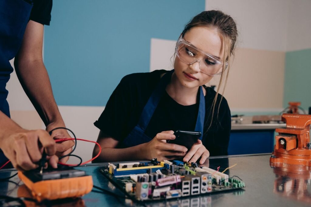 Female technician examining a circuit board in an electronics lab, using a smartphone for reference.